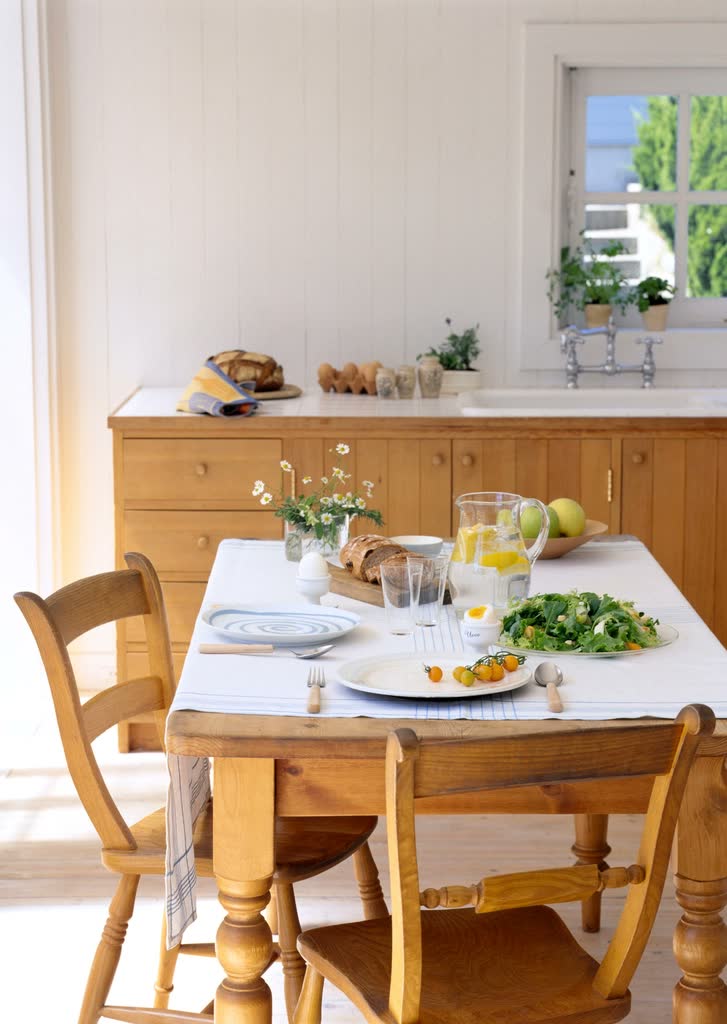 A honey pine dining table laid for a meal with chairs around it in a bright kitchen